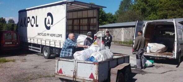 Mężczyźni ładują worki z odpadami do przyczepy i vana obok dużej ciężarówki z otwartą przestrzenią ładunkową. W tle widać drzewa i niebo z chmurami.