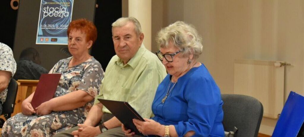 Grupa starszych osób siedzi na krzesłach w sali. Kobieta w niebieskiej sukience czyta z kartki. Obok niej mężczyzna w jasnej koszuli i kobieta w kwiecistej sukience. W tle plakat wydarzenia.