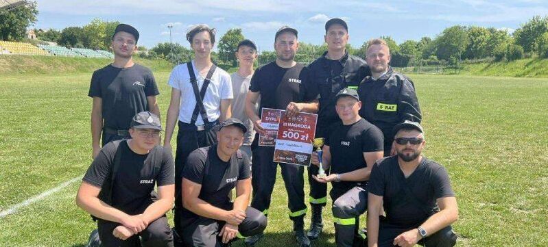 Grupa osób w czarnych koszulkach stoi i klęczy na zielonym boisku, uśmiechając się. Jeden z członków trzyma dużą tabliczkę z napisem "Nagroda 500 zł". W tle widać drzewa i część trybun.