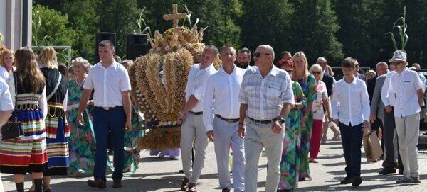 Uczestnicy procesji niosą dekoracje z kłosów zbóż zwieńczoną krzyżem. Ludzie ubrani są w tradycyjne i odświętne stroje na tle zielonych drzew. Atmosfera jest uroczysta i pogodna.