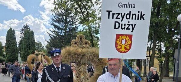 Parada na ulicy. Mężczyzna w koszuli niesie tabliczkę z napisem "Gmina Trzydnik Duży," obok niego osoba w stroju strażackim. Za nimi idzie grupa ludzi, niosąc ozdoby z siana. W tle drzewa i niebo z chmurami.