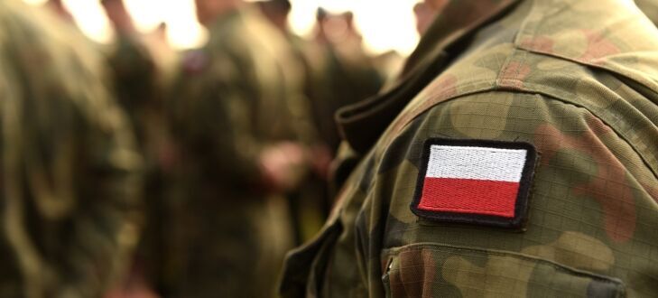 Na zdjęciu widać żołnierza w umundurowaniu z naszywką przedstawiającą polską flagę. W tle widoczni są nieostro inni żołnierze w podobnym umundurowaniu.