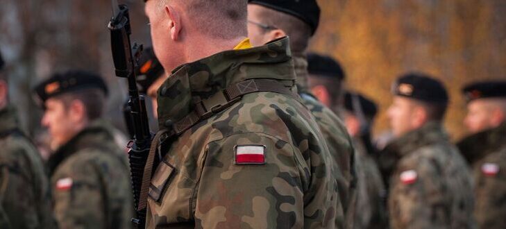 Grupa żołnierzy w mundurach moro stoi w szeregu. Na ramieniu jednego z żołnierzy widać naszywkę z polską flagą. Żołnierze mają czarne berety, a w tle widać rozmyte drzewa.