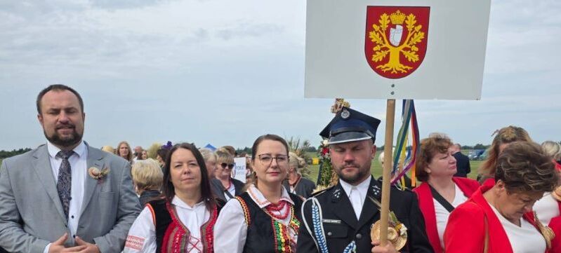 Grupa ludzi w tradycyjnych strojach oraz mundurze stoi na trawie, trzymają tablicę z napisem "Gmina Trzydnik Duży". Obok widoczny dekoracyjny wieniec dożynkowy.