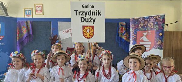 Dzieci w tradycyjnych strojach ludowych stoją w sali. Chłopcy mają białe koszule i kapelusze, dziewczynki kolorowe spódnice i wianki. Trzymają tablicę z napisem "Gmina Trzydnik Duży". W tle widać kolorowe dekoracje.