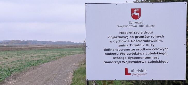 Na zdjęciu widać tablicę informacyjną dotyczącą modernizacji drogi dojazdowej do gruntów rolnych w gminie Trzydnik Duży. Obok widać nieutwardzoną drogę i otaczający krajobraz pól.