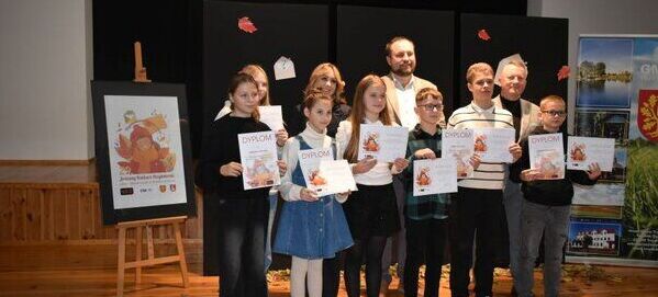 Grupa dzieci i dorosłych stoi na scenie, trzymając dyplomy. W tle plakat z jesiennym motywem. Drewniana podłoga, tablice i banery po bokach.