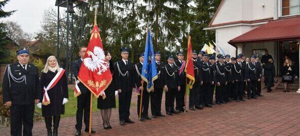 Grupa strażaków w galowych mundurach stoi w szeregu przed budynkiem, przy którym są drzewa iglaste. Kilka osób trzyma flagi w różnych kolorach, w tym biało-czerwoną z orłem.