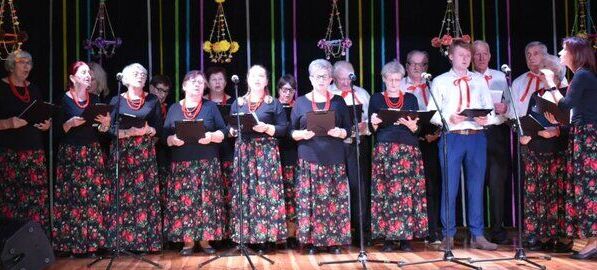 Grupa osób w tradycyjnych polskich strojach śpiewa na scenie, trzymając nuty. Tło ozdobione kolorowymi pionowymi tasiemkami i dekoracjami. Drewno na podłodze sugeruje lokalny występ lub festiwal.