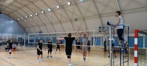 Grupa osób gra w siatkówkę na hali sportowej. Jeden z zawodników odbija piłkę przy siatce, sędzia obserwuje grę z podwyższenia. Poziome światła oświetlają przestronne wnętrze.