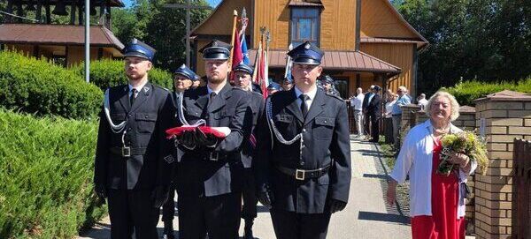 Grupa strażaków w mundurach maszeruje przed kościołem, niosąc flagę. Obok nich idzie kobieta w czerwonej spódnicy z bukietem kwiatów. W tle widać drewniany budynek i zebranych ludzi.