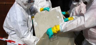 Osoby w ochronnych kombinezonach i rękawicach trzymają kawałek azbestu, obszar zabezpieczony białą i czerwoną taśmą.