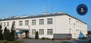 Budynek dwukondygnacyjny Szkoły Podoficerskiej Marynarki Wojennej z flagą Polski, tablicą informacyjną, antenami na dachu, pod niebieskim niebem.
