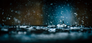 Krople deszczu uderzające w mokrą powierzchnię, tworzące rozpryski wody na ciemnym tle z rozmytym światłem w tle.