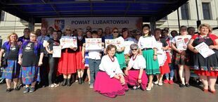 Grupa osób stoi na scenie na zewnątrz, trzymają certyfikaty; dwie dziewczynki w ludowych strojach siedzą z przodu; w tle baner "Powiat Lubartowski".