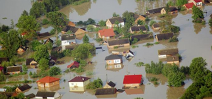 Zdjęcie przedstawia osiedle domów jednorodzinnych częściowo zatopionych przez powódź. Woda pokrywa ogrody i dociera do dachów, otaczając domostwa.