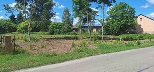 Droga asfaltowa prowadzi obok pustego, zarośniętego działki z kilkoma drzewkami. W tle widać budynek i niebieskie niebo z chmurami. Całość otoczona zielenią.