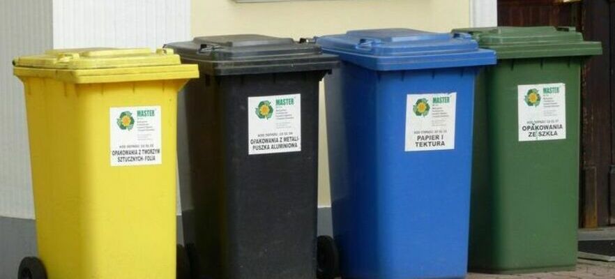 Cztery kolorowe pojemniki do segregacji odpadów ustawione obok siebie: żółty, czarny, niebieski i zielony, każdy z naklejką symbolizującą recykling.
