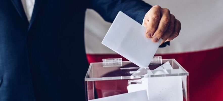 Osoba w czarnym garniturze wrzuca kartę do głosowania do przezroczystej urny wyborczej na tle czerwonej zasłony.