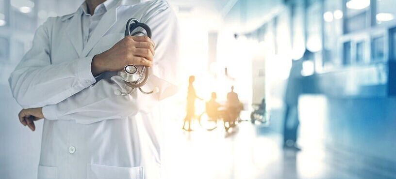 Lekarz w białym labcie trzymający stetoskop na przedzie, w tle rozmazany widok korytarza szpitalnego z sylwetkami pacjentów.