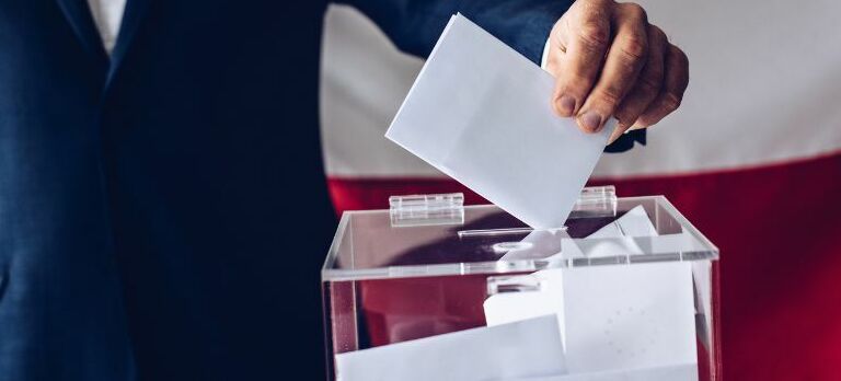 Osoba wrzuca białą kartkę do przezroczystej urny wyborczej na tle flagi.