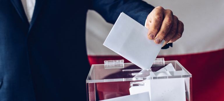 Osoba w garniturze wrzuca kartkę do przezroczystej urny wyborczej, na tle niewyraźnej flagi.