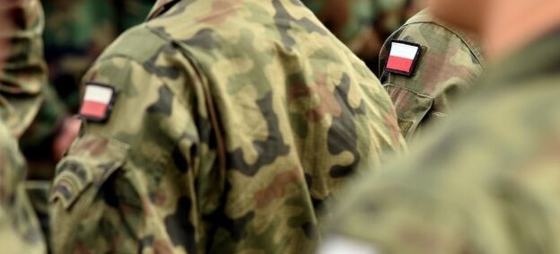 Żołnierze w mundurach w zielonym kamuflażu stoją ramię w ramię. Na ramionach znajdują się naszywki z polską flagą.