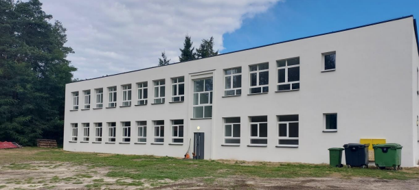 Dwupiętrowy biały budynek z wieloma oknami, ustawiony na tle drzew. Przed nim znajduje się trawnik oraz kilka pojemników na śmieci. Na niebie widać lekkie zachmurzenie.