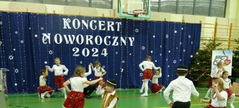 Grupa dzieci w tradycyjnych strojach wykonuje taniec ludowy na koncercie noworocznym w hali sportowej, z dekoracjami i napisem "KONCERT NOWOROCZNY 2024".