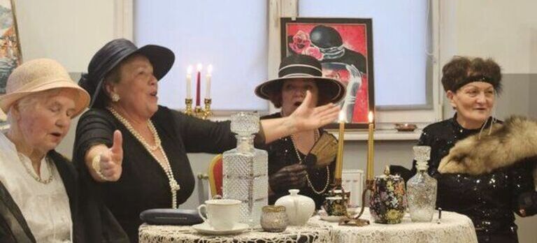Cztery starsze kobiety w kapeluszach ze stolikiem ze świecami, filiżankami i karafką w tle.