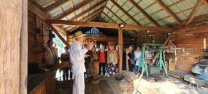 Grupa ludzi słucha przewodnika w tradycyjnym drewnianym młynie. Wnętrze jest rustykalne, z drewnianymi belkami i starymi narzędziami młyńskimi.