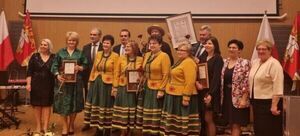 Grupa ludzi w żółtych szarfach stoi na scenie z certyfikatami, otoczona przez oficjele i flagi, podczas formalnej ceremonii.