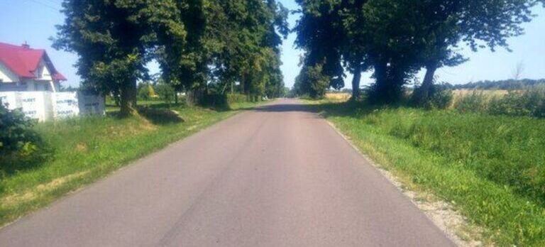 Asfaltowa droga prowadząca przez wiejski krajobraz z drzewami po obu stronach i domem po lewej. Ciepły, słoneczny dzień.