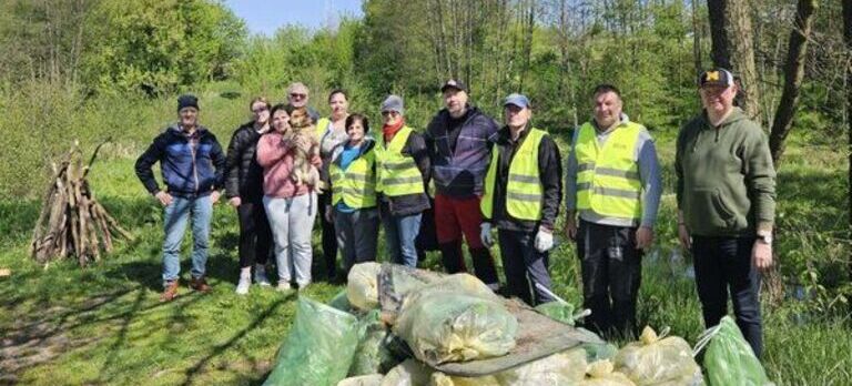 Grupa ludzi w kamizelkach odblaskowych stoi na tle lasu, z uśmiechami, obok skumulowanych worków z zebranymi odpadami.