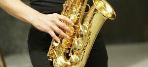 Osoba trzyma złoty saksofon altowy, na którym gra. Widoczne są szczegóły instrumentu i dłonie muzyka.