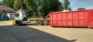 Ciężarówka z przyczepą obok dużej czerwonej przemysłowej kontenera na odpady, na tle zadrzewionego terenu i budynku.