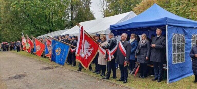 Grupa osób stoi przydrożnie z flagami i sztandarami obok namiotów w parku lub leśnej alei.