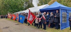 Grupa osób stoi przydrożnie z flagami i sztandarami obok namiotów w parku lub leśnej alei.