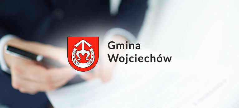 Osoba trzyma długopis nad dokumentem. Po lewej stronie znajduje się herb gminy Wojciechów: czerwony tarcza z białym motywem serca i kotwicy. Obok widnieje napis "Gmina Wojciechów".