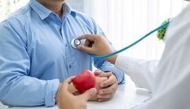 Osoba w białym fartuchu używa stetoskopu do badania innego człowieka, który trzyma czerwone serce w dłoniach, symbolizujące zdrowie.