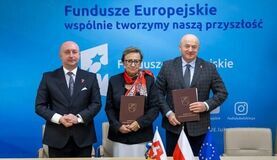 Trzy osoby stoją przy stole z dokumentami i flagami Polski oraz UE, trzymają teki. Baner w tle: "Fundusze Europejskie - wspólnie kształtujemy naszą przyszłość".