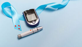 Na jasnoniebieskim tle leży niebieska wstążka z trzema czerwonymi serduszkami, glukometr oraz biały z szarymi elementami lancet do pobierania krwi. Wstążka symbolizuje świadomość cukrzycy.