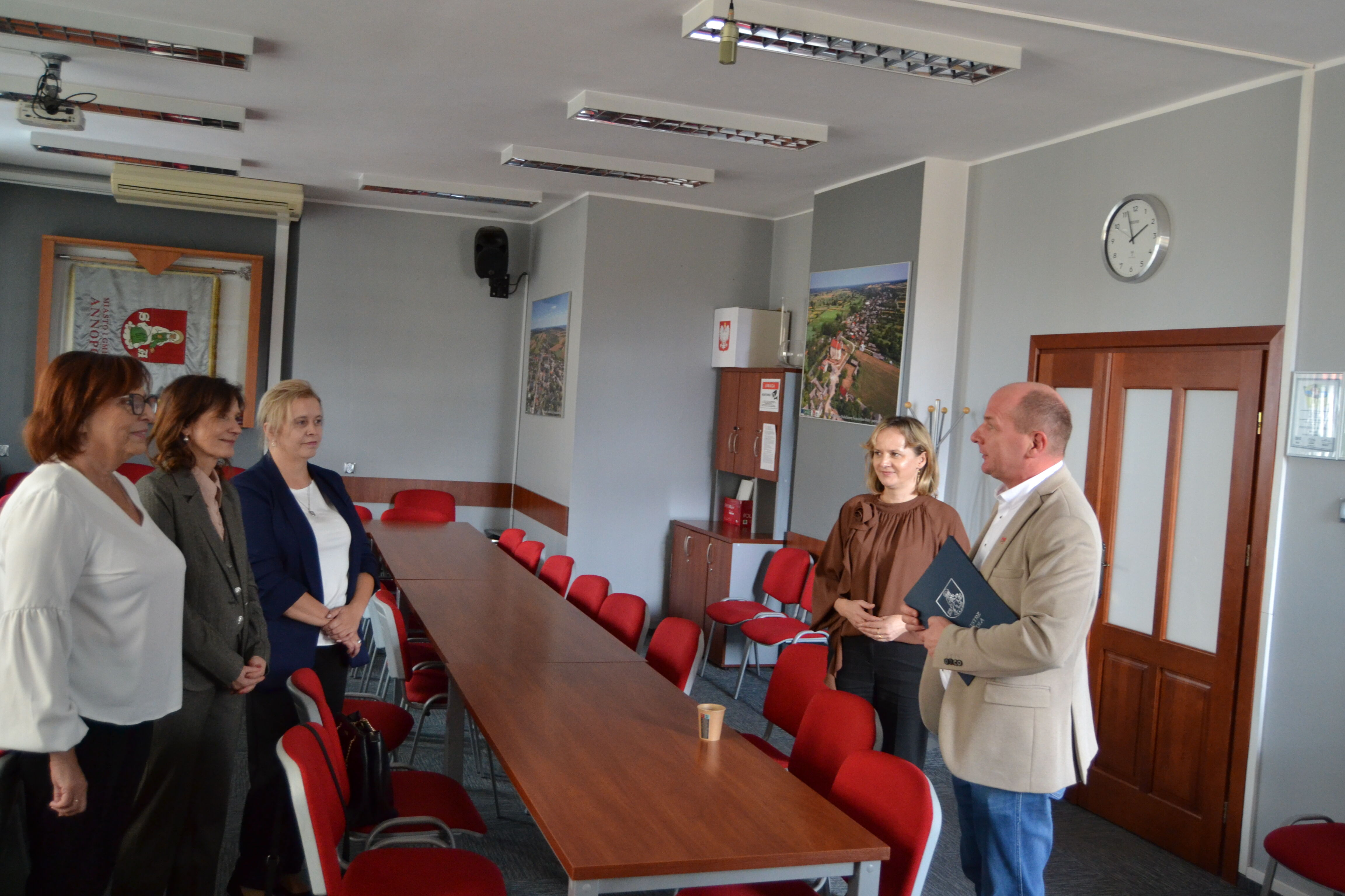 Na zdjęciu widać grupę osób stojących w sali konferencyjnej z czerwonymi krzesłami i długim stołem. Jedna osoba trzyma teczkę. W tle widać zegar na ścianie i różne dekoracje.