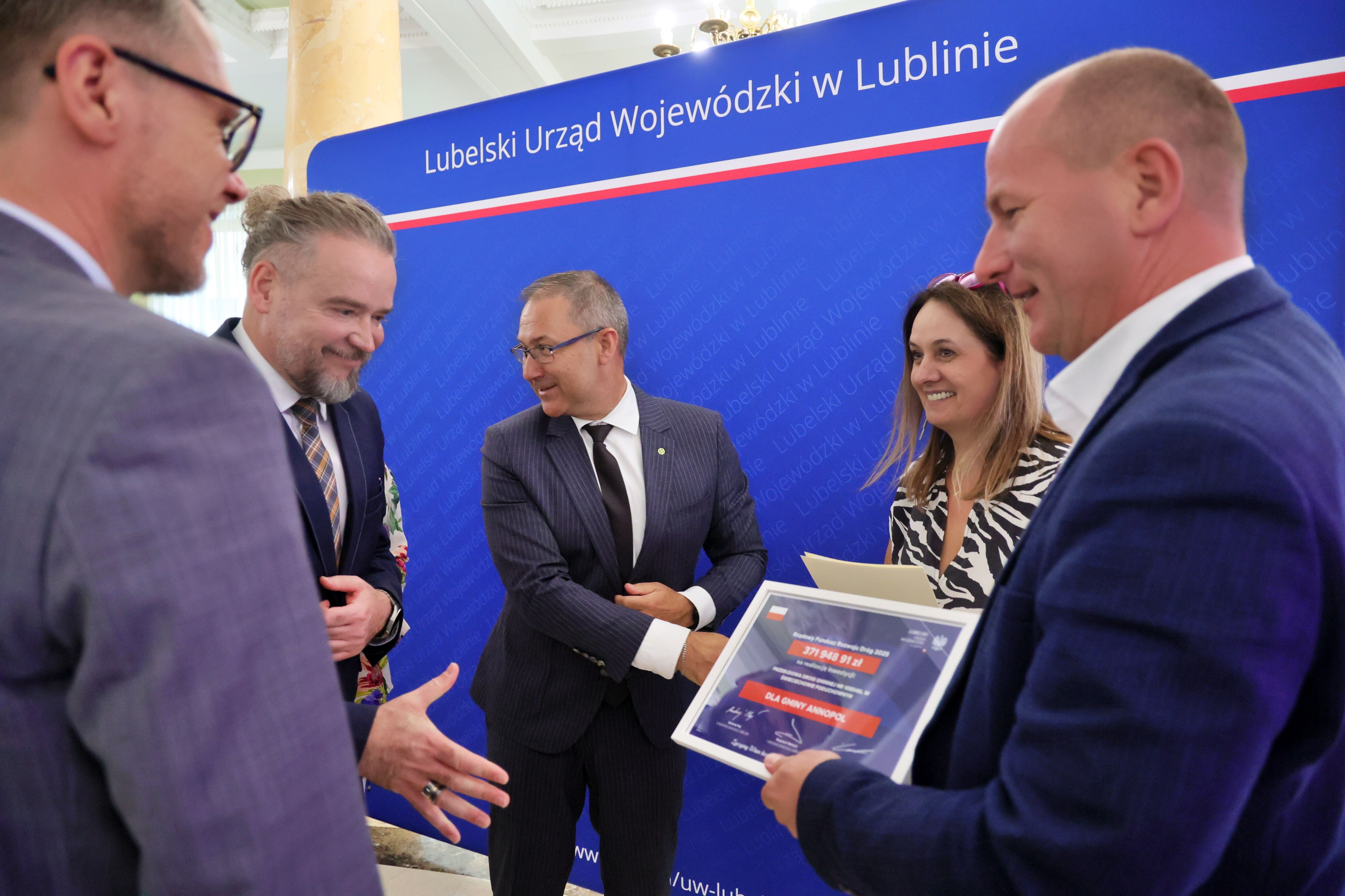 Grupa osób w eleganckich strojach rozmawia przed niebieskim tłem z napisem "Lubelski Urząd Wojewódzki w Lublinie." Jedna osoba trzyma certyfikat lub dyplom. Wszyscy wyglądają na zadowolonych.
