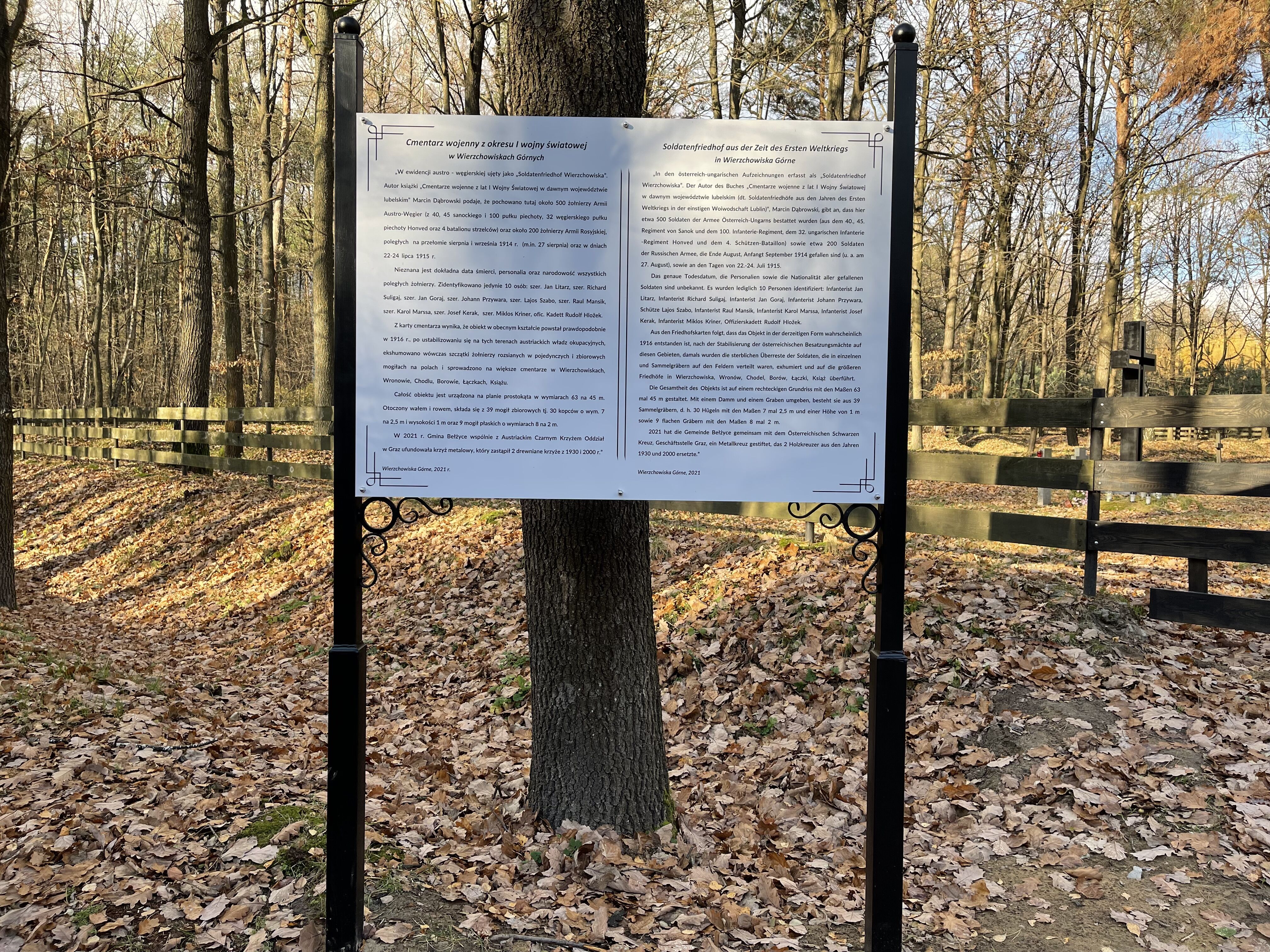 Zdjęcie przedstawia białą tablicę z informacjami historycznymi o cmentarzu z I Wojny Światowej w Wierzchowiskach. W dalszym planie widać drzewa, a na ziemi znajdują się opadłe liście z drzew.