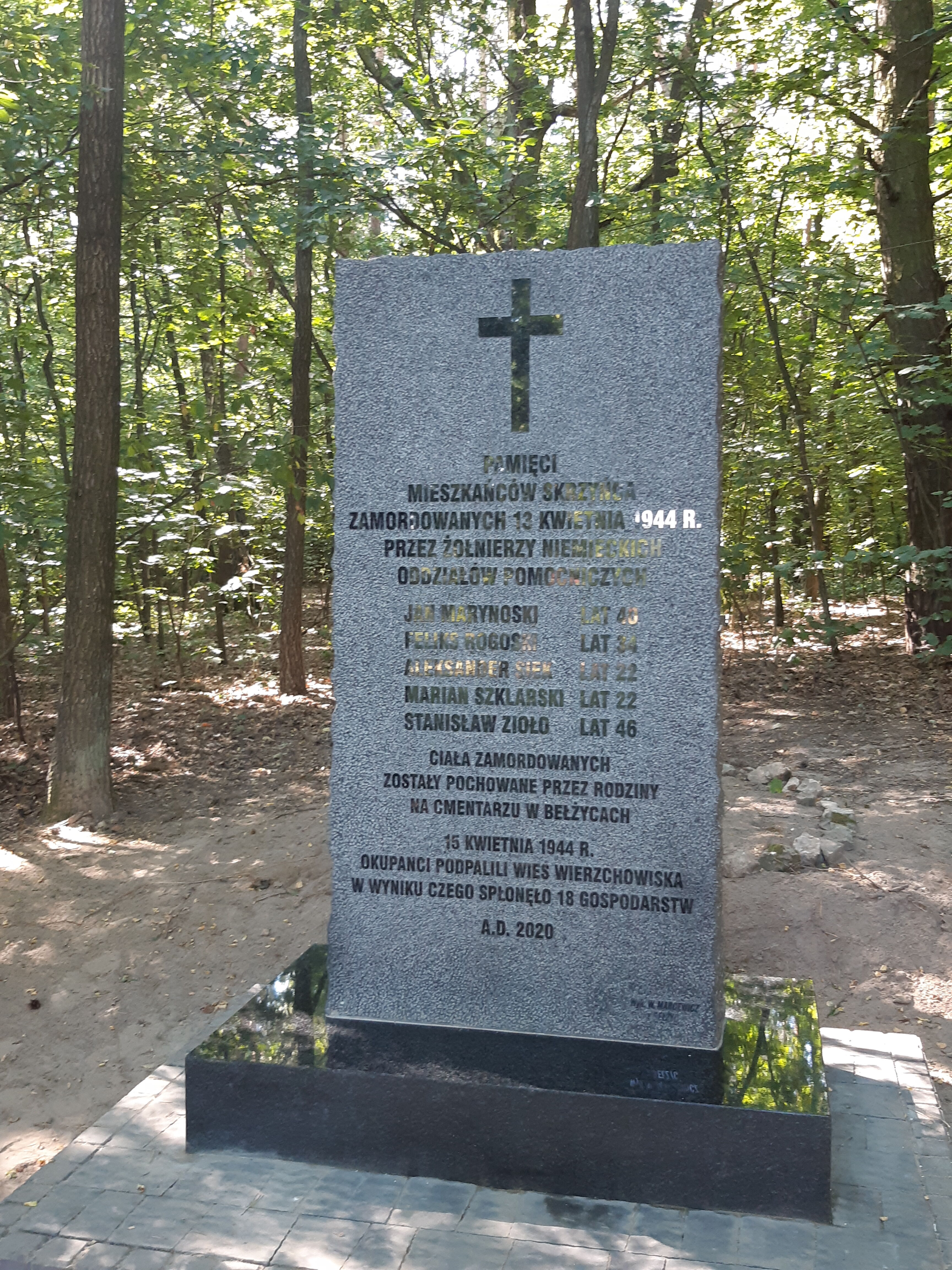 Grafika przedstawia szary pomnik upamiętniający wydarzenia z dnia 13 kwietnia 1944 r. W drugim tle zielony las.