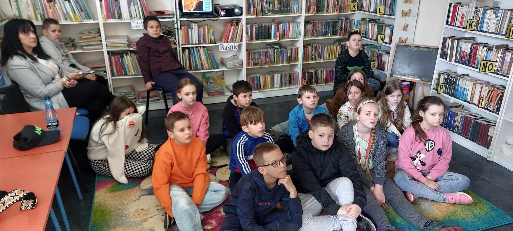Grupa dzieci siedzi na podłodze w bibliotece, skupiając uwagę na przedniej części pomieszczenia. Po lewej stronie siedzi dorosła osoba.