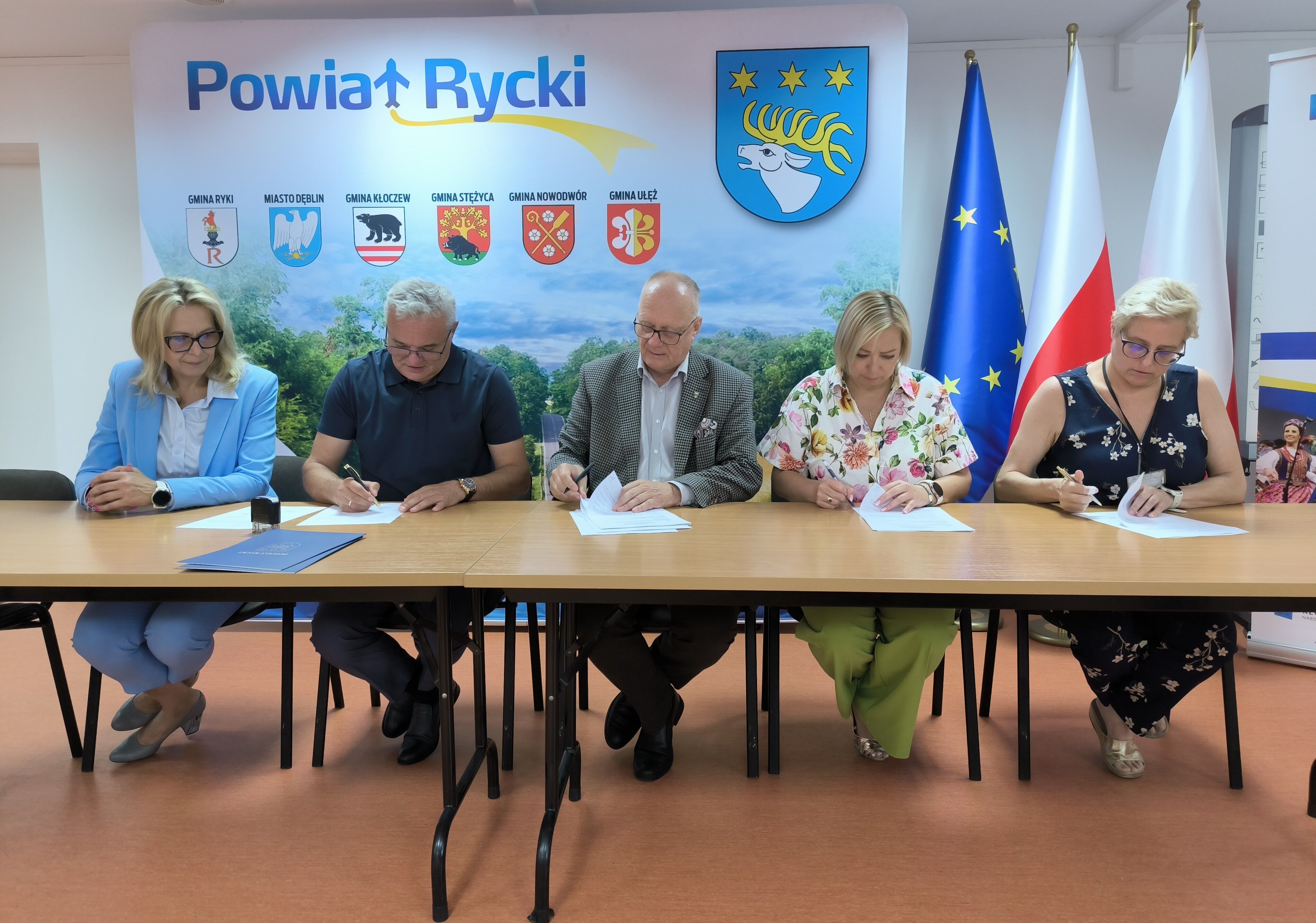Pięć osób siedzi przy stole, podpisując dokumenty. Za nimi widoczna jest tablica z nazwami gmin i herbami, a także flagi Polski i Unii Europejskiej. W tle znajduje się plakat z napisem "Powiat Rycki".