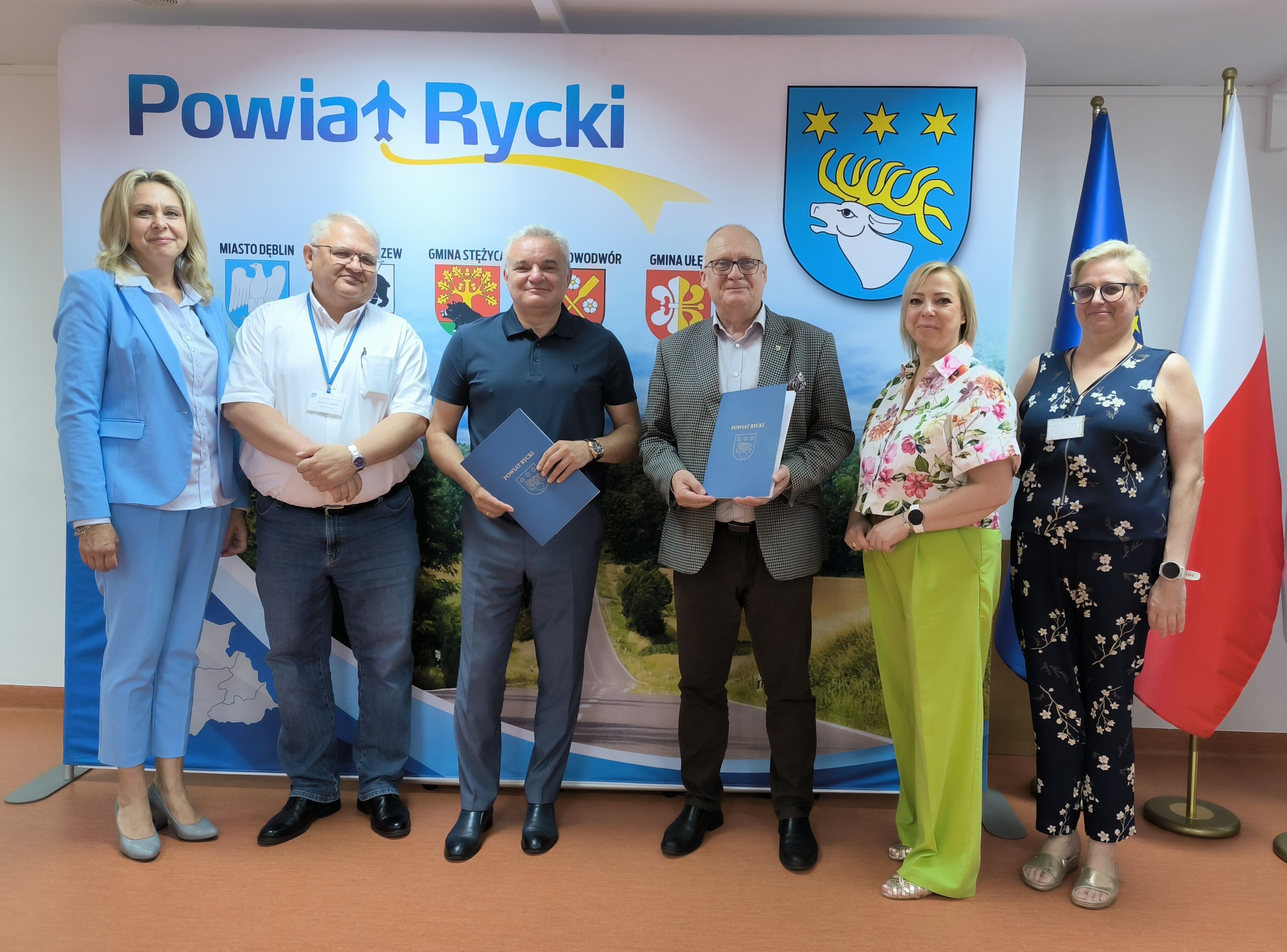 Sześć osób stoi przed banerem z logo Powiatu Ryckiego. Dwie osoby trzymają dokumenty. W tle widoczne są flagi Polski i powiatu oraz zdjęcie krajobrazu. Grupa uśmiecha się do zdjęcia.