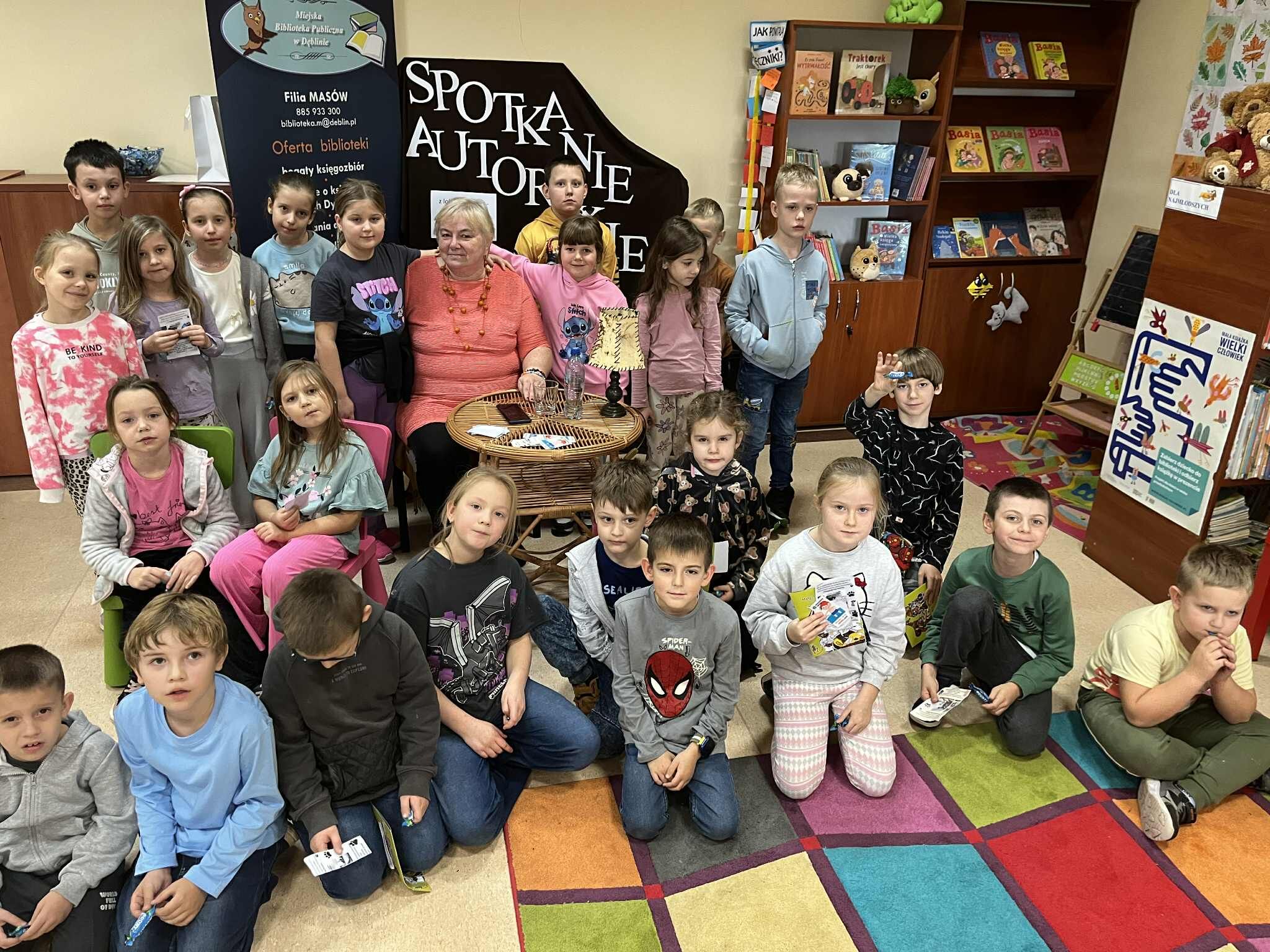 Grupa dzieci i dorosła osoba siedzą w bibliotece na kolorowym dywanie. Dookoła widać książki na półkach oraz plakat promujący spotkanie autorskie. Dzieci większość patrzy w stronę aparatu.
